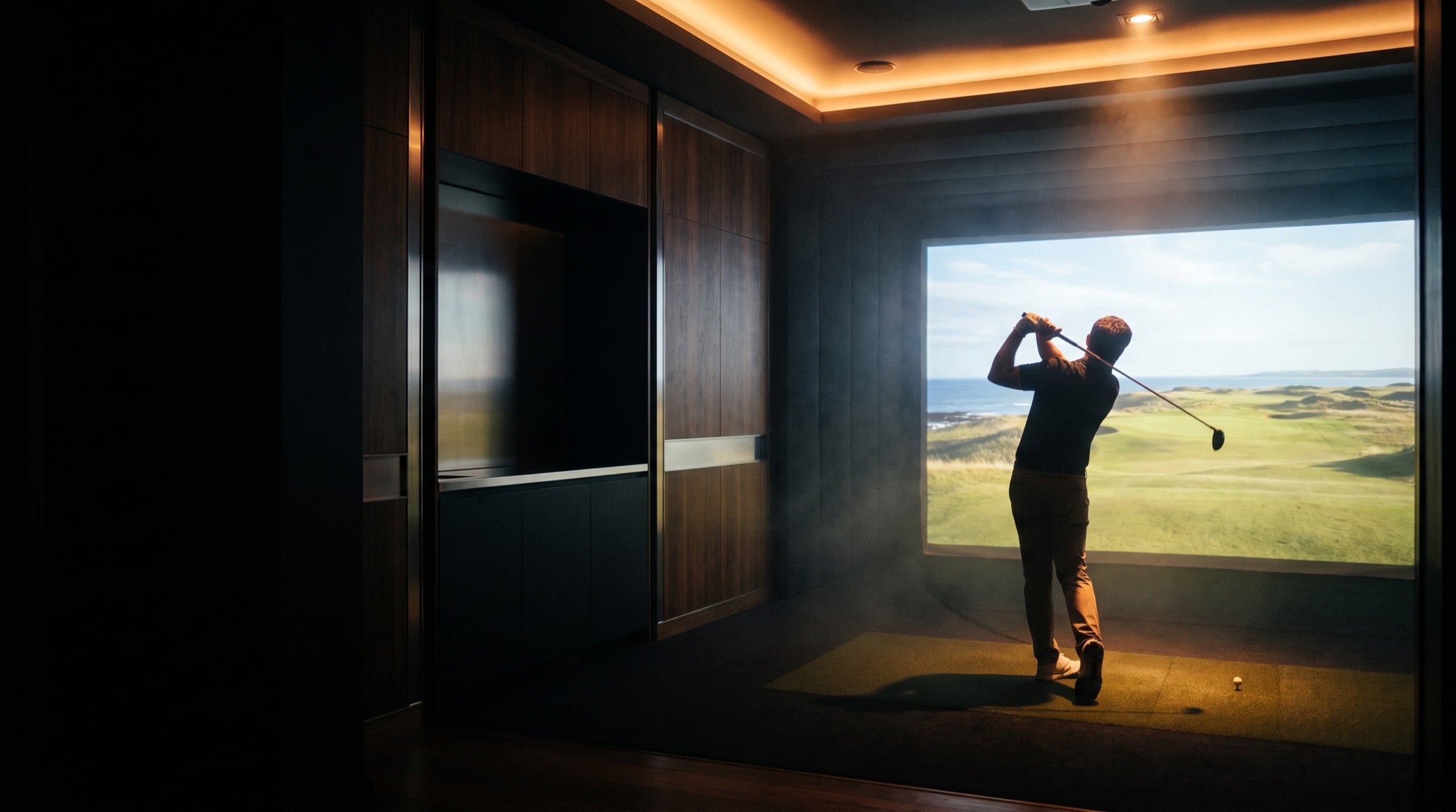 A golfer mid-swing in a darkened indoor golf simulator bay, facing a lit screen showing a links course.