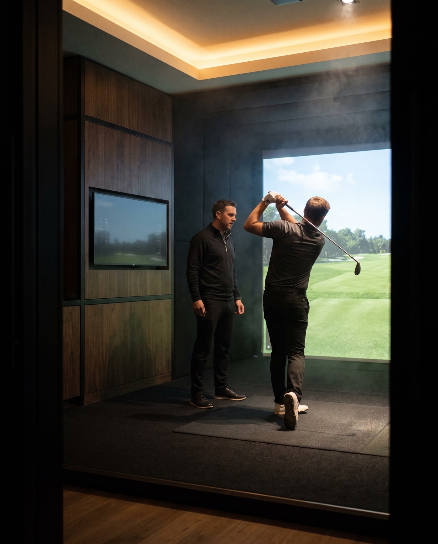 A golf coach watching a golfer complete a swing inside an indoor simulator bay.