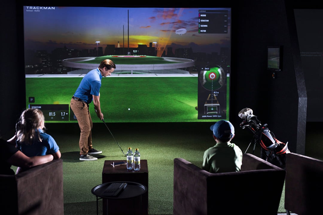 A group seated in a simulator bay watching one player hit toward a target.