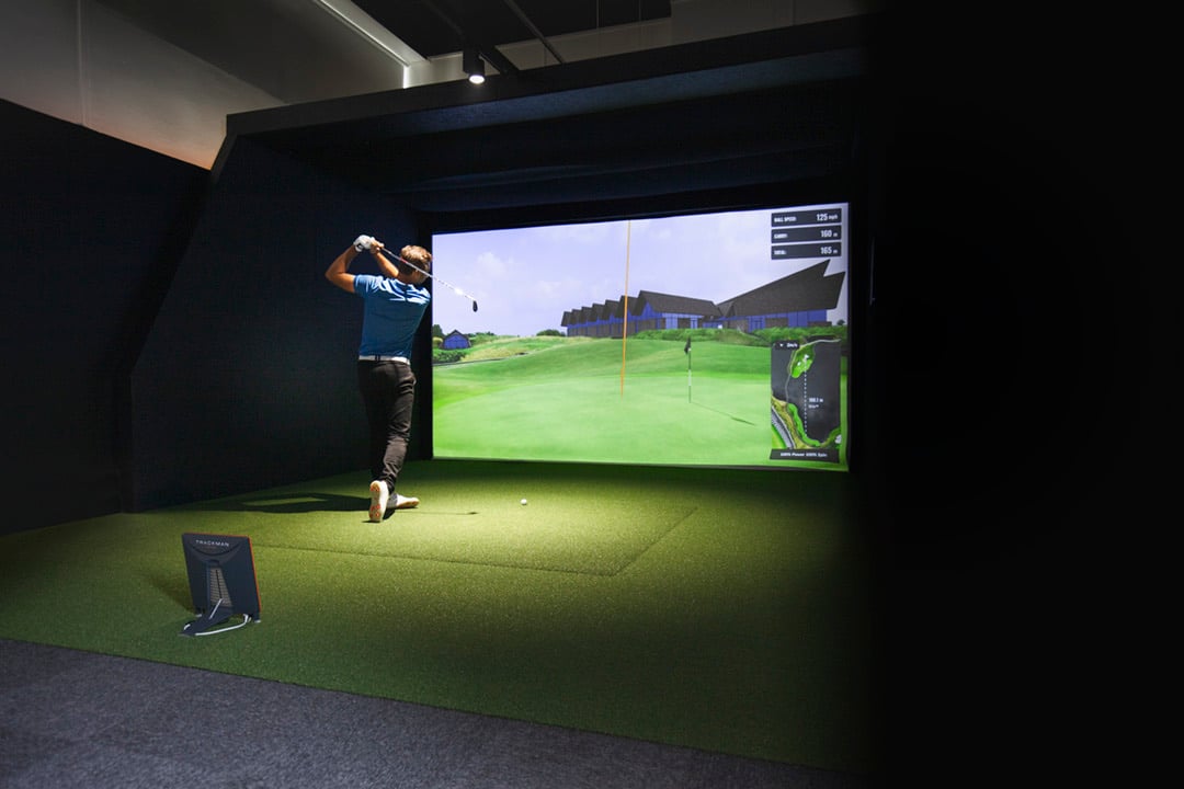 A golfer mid-swing in a simulator bay, with shot data displayed on the screen.